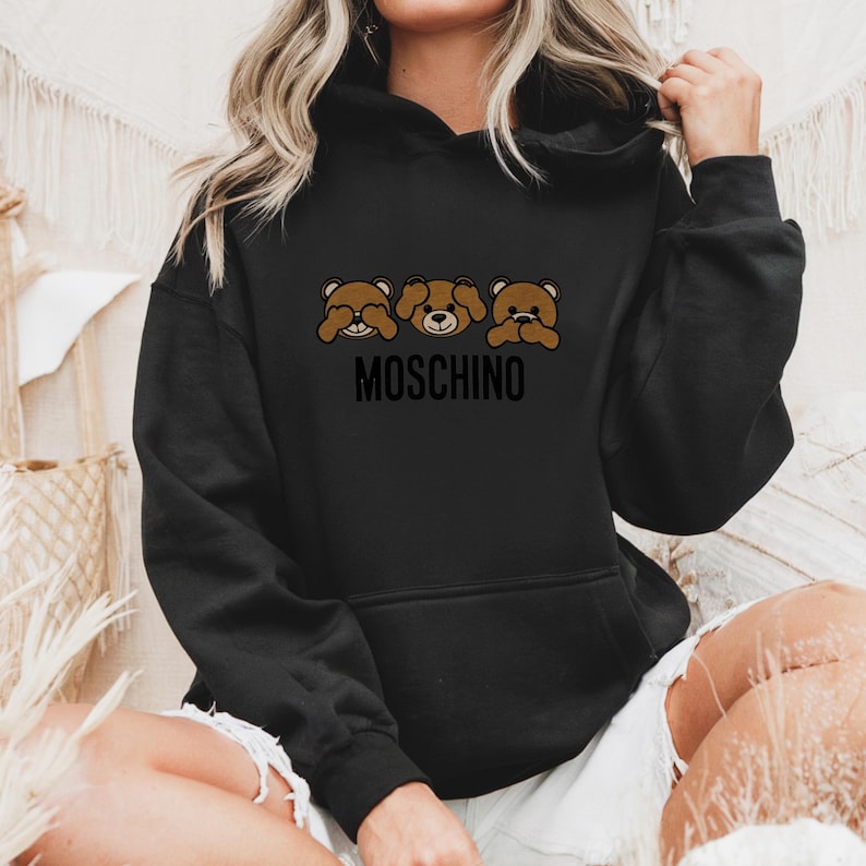May include: Black hoodie with a graphic of three brown teddy bears covering their eyes, mouth, and ears. The text "MOSCHINO" is printed below the bears.