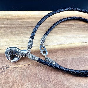 Könnte beinhalten: Eine schwarze Lederkette mit einem silbernen Boxhandschuh-Anhänger. Die Kette ist geflochten und hat einen silbernen Verschluss.