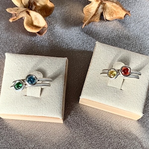 Puede incluir: Tres anillos de plata con piedras preciosas de colores. Los anillos se muestran en una caja acolchada blanca. Las piedras preciosas son verdes, azules y naranjas.