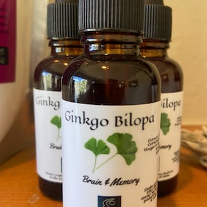 Puede incluir: Tres botellas de vidrio marrón con goteros, etiquetadas con "Ginkgo Bilopa" y "Brain & Memory". Las botellas están llenas de un líquido oscuro y tienen etiquetas blancas con texto negro. La etiqueta también presenta un dibujo de una hoja de ginkgo.