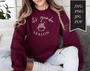 Puede incluir: Una sudadera color burdeos con el texto "it's gumbo season" en letras blancas. El texto va acompañado de una ilustración sencilla de una olla de gumbo con vapor saliendo de ella.
