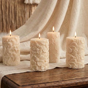May include: Four ivory pillar candles with floral relief patterns are lit, creating a warm glow. The candles are arranged on a wooden surface with a neutral linen cloth, creating a cosy, decorative ambiance.