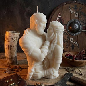 Puede incluir: Una vela de color crema que representa a una pareja vikinga abrazándose. La vela está intrincadamente esculpida con detalles de su ropa y rasgos. Otros elementos incluyen un escudo de madera, un cuerno para beber y un hacha pequeña.