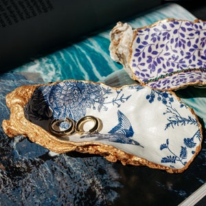 Może przedstawiać: Dwa ozdobne talerze w kształcie muszli ostrygi z złotymi akcentami. Jeden talerz jest biały z niebieskimi wzorami kwiatowymi i ptakiem, a drugi jest biały z fioletowymi wzorami kwiatowymi. Oba talerze trzymają pierścienie.