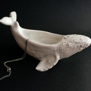 Puede incluir: Escultura de ballena de cerámica blanca con una superficie texturizada, diseñada como un plato para joyas. Una cadena plateada está drapeada sobre la espalda de la ballena.