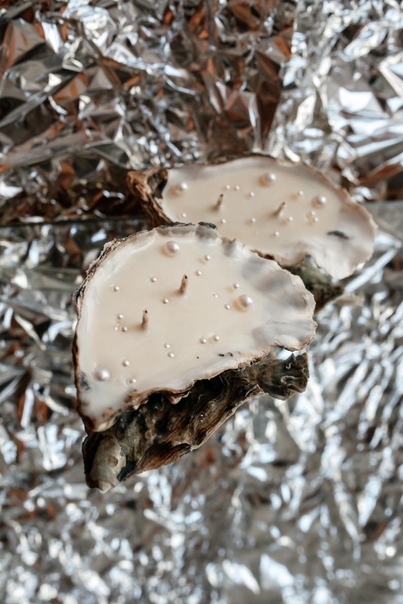 Two oyster shell candles with pearl decorations on a shiny silver foil background.