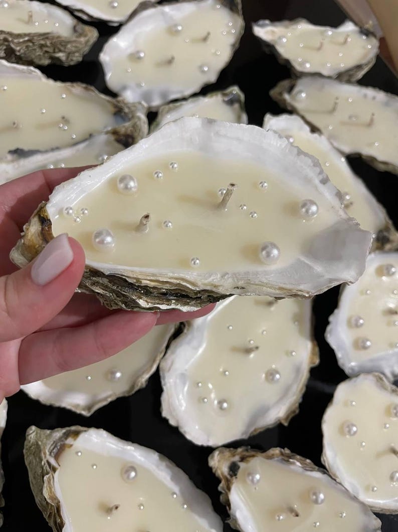 A woman's hand holds a large oyster shell candle with two wicks and decorative pearls. In the background, many similar candles are visible.