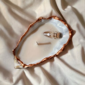 Könnte beinhalten: Eine weiße Kerze in einer goldgeränderten Austernschalen mit einem kleinen Glasfläschchen mit einem Korkstopfen.