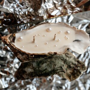 A single handmade candle in an oyster shell with two wicks and a scattering of pearls. It lies on a mirror reflecting silvery crumpled foil.