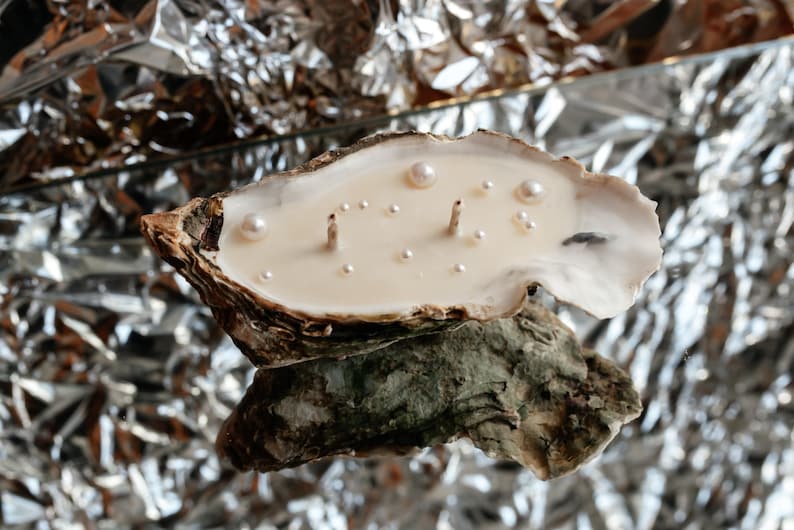 A single handmade candle in an oyster shell with two wicks and a scattering of pearls. It lies on a mirror reflecting silvery crumpled foil.
