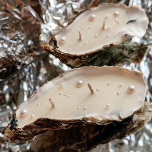 Vertical photo. Two large oyster candles featuring two wicks and faux pearls resting on a mirror that vividly reflects crumpled aluminum foil.