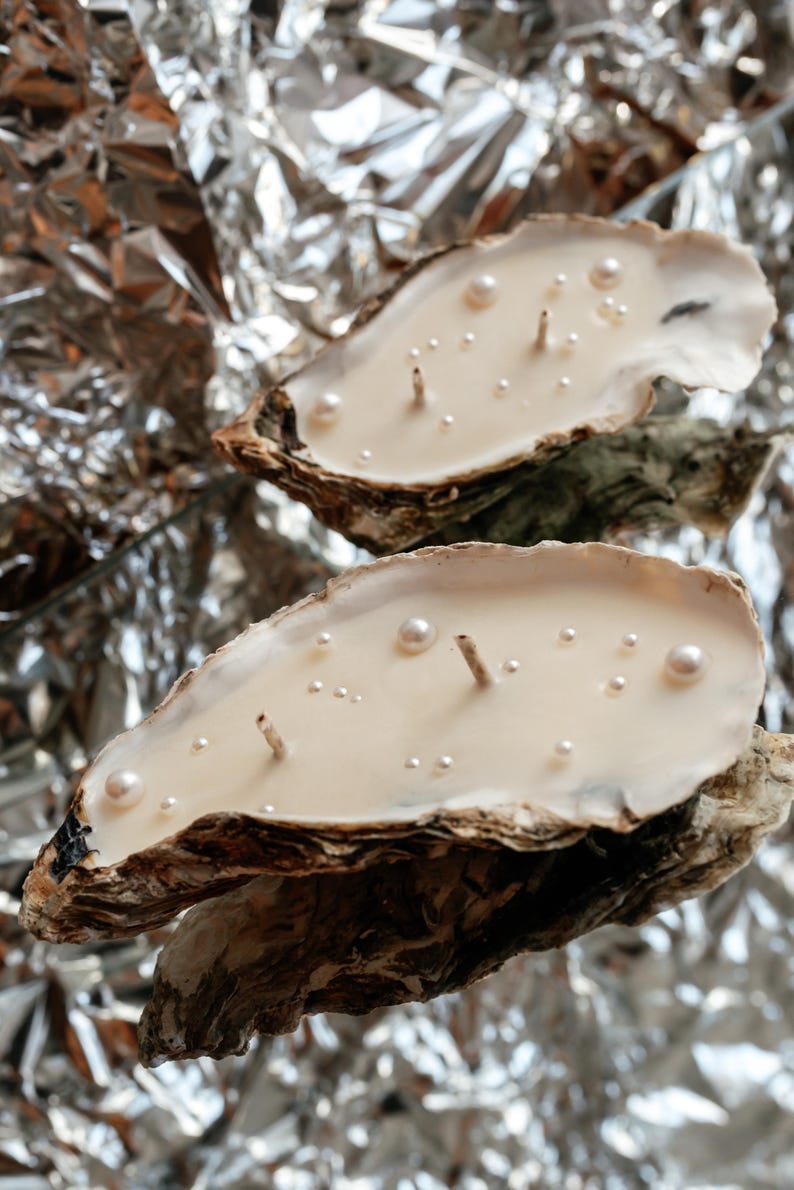 Vertical photo. Two large oyster candles featuring two wicks and faux pearls resting on a mirror that vividly reflects crumpled aluminum foil.