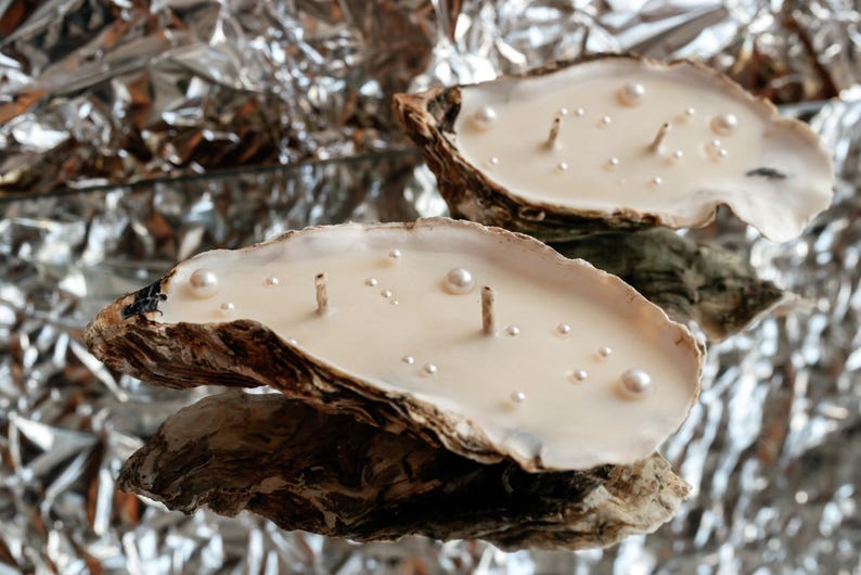 Two oyster shell candles with pearl decorations on a shiny silver foil background.