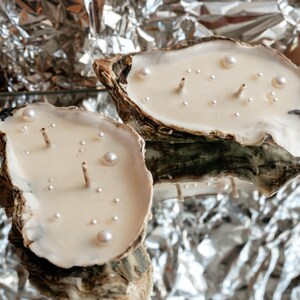 Two oyster candles with two wicks and white pearls. They are arranged horizontally on a mirror, creating an optical illusion by reflecting crumpled foil.