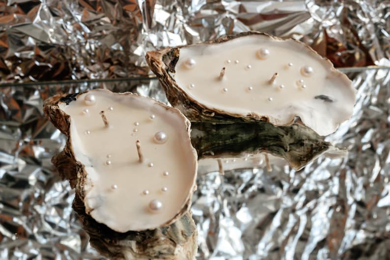 Two oyster candles with two wicks and white pearls. They are arranged horizontally on a mirror, creating an optical illusion by reflecting crumpled foil.