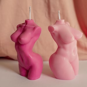 Puede incluir: Dos velas con forma de torso. Una es de color rosa intenso, la otra rosa claro. Ambas tienen una mecha en la parte superior. Las velas están sobre una superficie blanca con un fondo de tela rosa. Las velas son un artículo decorativo.