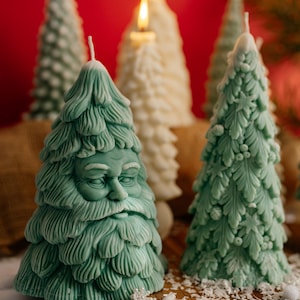 Puede incluir: Varias velas con forma de árbol de Navidad en tonos verdes y blancos, algunas encendidas. Una vela tiene la cara de Papá Noel. Las velas están dispuestas sobre una superficie de madera con adornos festivos.