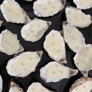 Top-down view of a large batch of finished double-wick oyster candles with pearls. They are densely and randomly laid out on a matte black background.