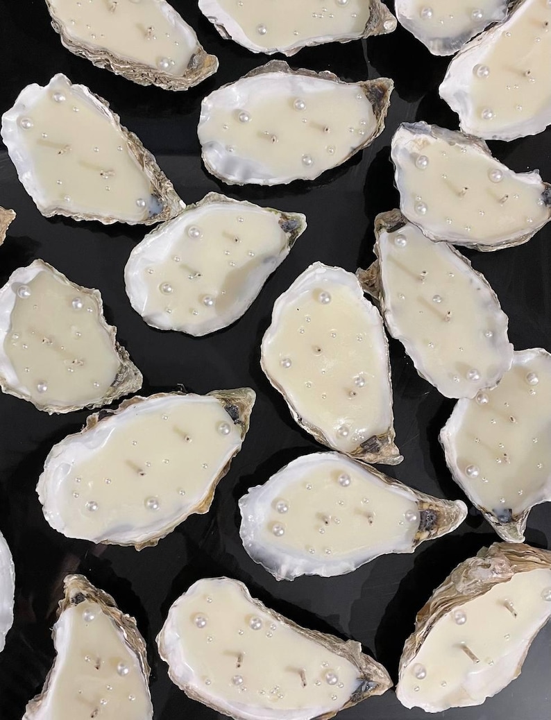 Top-down view of a large batch of finished double-wick oyster candles with pearls. They are densely and randomly laid out on a matte black background.