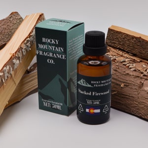 May include: A brown glass bottle of "Stacked Firewood" fragrance essential oil, 50ml, with a black cap, next to a matching box. The box and label feature the "Rocky Mountain Fragrance Co." logo and text. Wood pieces are in the background.