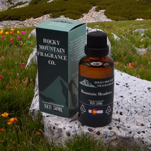 May include: A brown glass bottle and a matching box of Rocky Mountain Fragrance Co. essential oil, labeled "Mountain Meadows." The box and bottle have a black label with a mountain graphic and the Colorado flag. The net volume is 50ml.