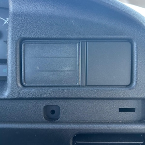 Puede incluir: Primer plano de un salpicadero de coche negro. El salpicadero presenta un compartimento rectangular con dos secciones, una abertura circular y una rejilla de ventilación rectangular. La superficie tiene una textura.