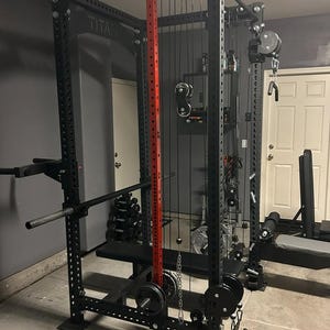 Könnte beinhalten: Ein schwarzes Krafttrainings-Power-Rack mit roter vertikaler Stütze. Das Rack umfasst eine Klimmzugstange, eine Gewichtsablage und Kabelzugmaschinen-Aufsätze. Gewichte und eine Bank sind ebenfalls sichtbar.
