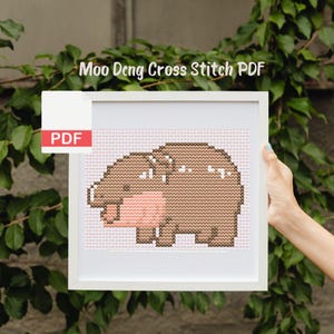 May include: A white frame with a PDF label in the top left corner. The frame contains a cross stitch pattern of a brown and pink pig. The title "Moo Deng Cross Stitch PDF" is at the top of the frame.