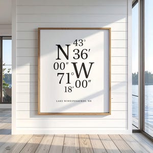 May include: A framed print displaying the coordinates 43° N 36' 00" W 71° 00' 18" and the text "LAKE WINNIPESAUKEE, NH". The print is in a wooden frame and is mounted on a white panelled wall. The room has a view of a lake.