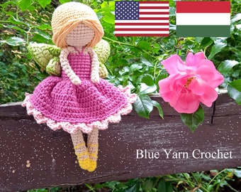 Puede incluir: Una muñeca de hada de crochet con un vestido rosa y alas verdes sentada en una valla de madera. La muñeca tiene el pelo rubio y zapatos amarillos. El fondo es un arbusto verde con una rosa rosa.