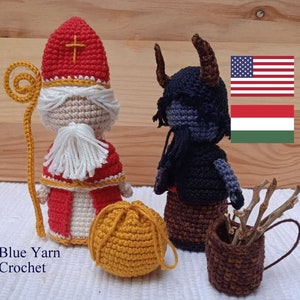 Pode incluir: Figuras de crochê de São Nicolau e Krampus, figuras tradicionais associadas ao Natal em alguns países europeus. São Nicolau segura um cajado dourado e um saco, enquanto Krampus segura uma cesta de ramos. As figuras estão em pé sobre uma superfície de madeira com bandeiras americana e húngara no fundo. O texto "Blue Yarn Crochet" é visível no canto inferior esquerdo.