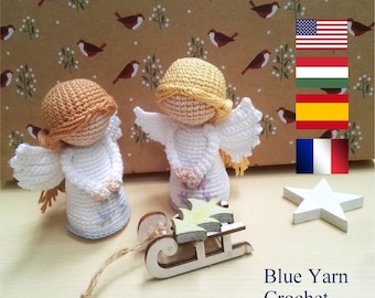 May include: Two crocheted angels with white dresses and wings, one with blonde hair and the other with brown hair. The angels are sitting on a wooden sled with a green and yellow Christmas tree. The background is a brown paper with a white pattern of birds and flowers. The text "Blue Yarn Crochet" is at the bottom of the image.