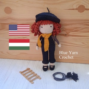 Może przedstawiać: Lalka szydełkowa z czerwonymi włosami, w czarnym berecie, żółtym szaliku i czarnych ogrodniczkach. Lalka stoi na drewnianej powierzchni z małą drewnianą drabiną i sznurkiem szydełkowym w kolorze szarym z czarnymi frędzlami. W obrazie widoczny jest tekst "Blue Yarn Crochet".