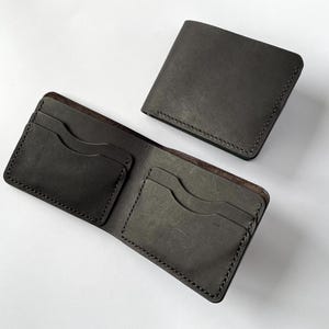 Pode incluir: Duas carteiras de couro cinza escuro são exibidas em uma superfície branca. Uma carteira está aberta, revelando ranhuras para cartões, enquanto a outra está fechada. Ambas as carteiras têm costuras visíveis nas bordas, mostrando o artesanato.