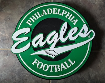 Puede incluir: Un letrero circular verde y blanco con las palabras "Philadelphia Eagles Football" en una fuente blanca cursiva. El letrero tiene un fondo negro con un diseño de ala de águila blanca.