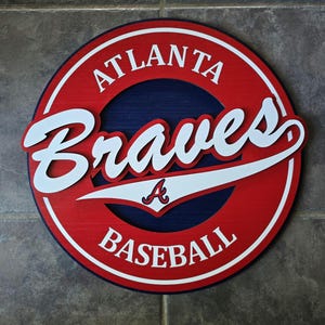 May include: A round wooden sign featuring the Atlanta Braves baseball team logo. The sign has a red outer ring with the words "ATLANTA BASEBALL" in white. The word "Braves" is in white script over a blue background.