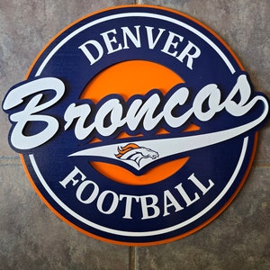 May include: A round wooden sign with a navy blue background and an orange center. The sign says "Denver Broncos Football" in white lettering with a stylized bronco logo in the center.