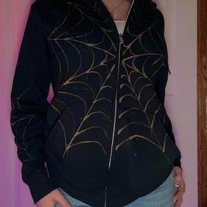 May include: A black zip-up hoodie with a gold spiderweb design. The hoodie has a hood and long sleeves.