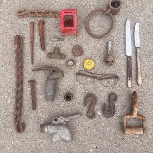 May include: A collection of assorted metal objects including rusted chains, a red metal box, a large ring, two knives, a brass T-fitting, a yellow bottle cap, eyeglasses, two metal hooks, a handle, and two metal spigots arranged on a gray surface.