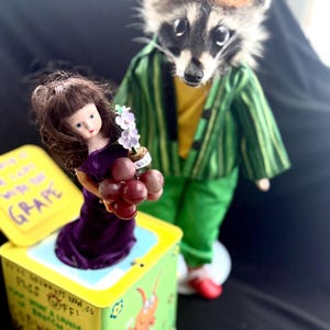 May include: A whimsical scene featuring a raccoon dressed in a green striped jacket and pants, standing behind a doll holding grapes and flowers. The doll emerges from a yellow box with text and illustrations.