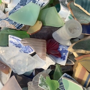May include: A collection of sea glass and pottery shards in various colors and shapes. Pieces include translucent green, white, and blue glass, along with fragments of patterned ceramic plates and cups. The assortment creates a textured, colorful display.