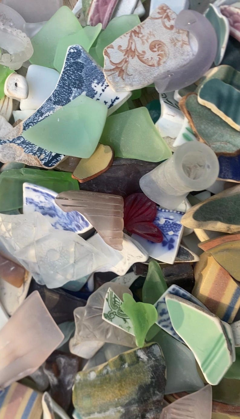 May include: A collection of sea glass and pottery shards in various colors and shapes. Pieces include translucent green, white, and blue glass, along with fragments of patterned ceramic plates and cups. The assortment creates a textured, colorful display.