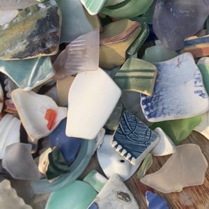 May include: A collection of sea glass and pottery shards in various colors, including white, blue, green, and yellow. The pieces have smooth, rounded edges and are of different shapes and sizes. The image shows a close-up view of the fragments.
