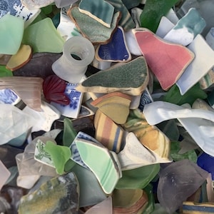 May include: A collection of sea glass and ceramic shards in various colors, including green, blue, pink, and white. Some pieces have painted patterns and designs. The assortment appears to be a mix of broken pottery and smoothed glass.