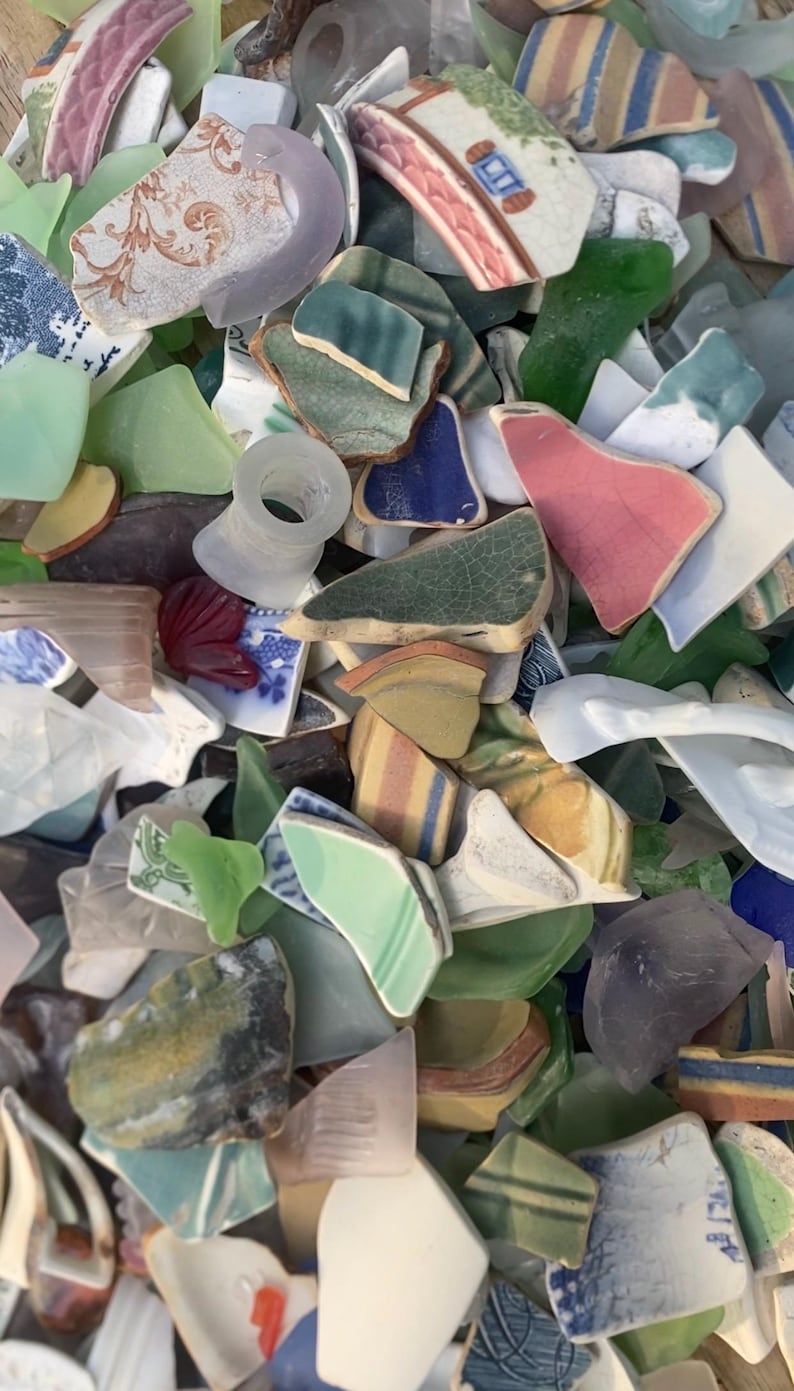 May include: A collection of sea glass and ceramic shards in various colors, including green, blue, pink, and white. Some pieces have painted patterns and designs. The assortment appears to be a mix of broken pottery and smoothed glass.