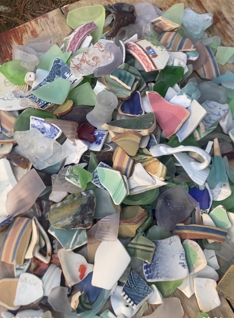 May include: A collection of sea glass and pottery shards in various colors, including green, blue, pink, and white. The fragments have different patterns and textures, creating a mosaic-like appearance. The pieces are of varying sizes and shapes.