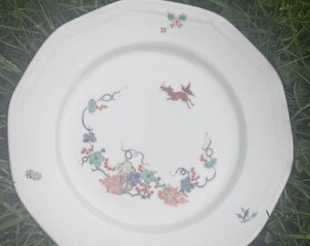 May include: A white octagonal ceramic plate featuring a delicate hand-painted design. The center displays a small red leaping deer surrounded by stylized floral motifs in shades of green, blue, and red. Additional small floral accents are placed.