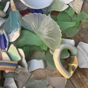 May include: A collection of sea glass and ceramic shards in various shapes and colors, including white, green, blue, and beige. Some pieces have decorative patterns. The items are arranged on a wooden surface.