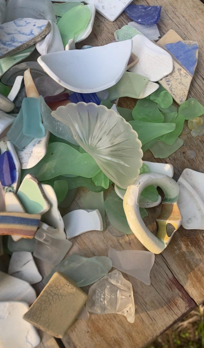 May include: A collection of sea glass and ceramic shards in various shapes and colors, including white, green, blue, and beige. Some pieces have decorative patterns. The items are arranged on a wooden surface.