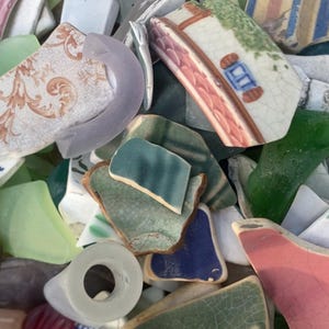 May include: A collection of sea glass and pottery shards in various colors. Pieces include green, blue, pink, and white fragments, some with painted designs. The assortment suggests a craft or decorative theme, perfect for mosaics or jewelry.